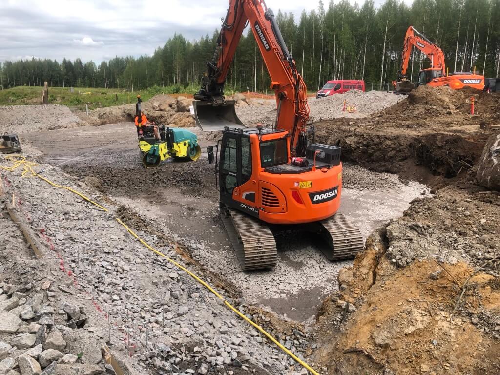 Kaksi oranssia Doosan-kaivinkonetta ja yksi henkilön ohjaama murskaaja työmaalla metsätaustan vieressä.